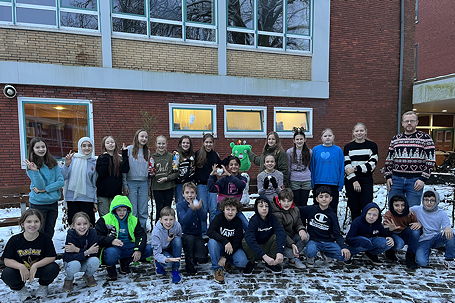 Das Foto zeigt die Klasse 6a mit ihrem Schneemann auf dem Pausenhof des TGG mit ihrem grünen Klassenmaskottchen (Dezember 2023).