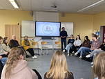 Das Foto zeigt einen Blick in den Klassenraum mit dem Referenten und der Referentin; die Schülerinnen und Schüler sitzen im Kreis; auf der digitalen Tafel ist im Hintergrund das Logo der Veranstaltung zu sehen.